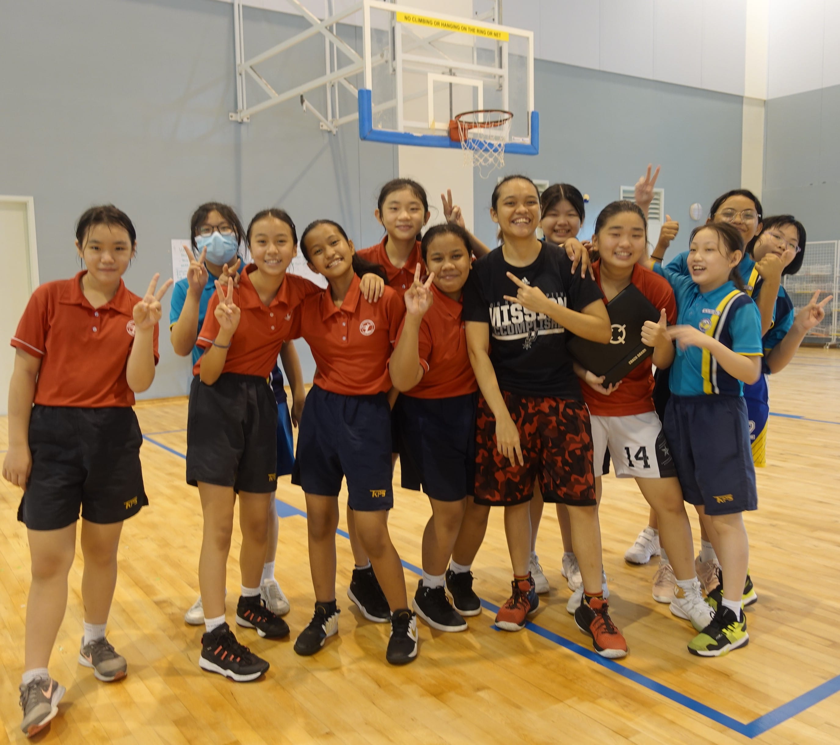 A group of young girls in red and teal uniforms pose with peace signs on an indoor basketball court.